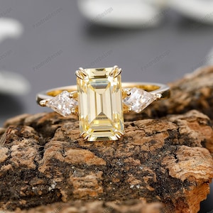 May include: A gold engagement ring with a large, rectangular, yellow gemstone and two smaller, triangular, clear gemstones on either side.