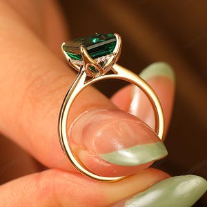 Può includere: Un anello d'oro con una grande pietra verde taglio smeraldo. L'anello ha una fascia sottile e piccole pietre di accento attorno alla gemma principale. L'anello è tenuto in alto, mostrando l'intricato design e la brillantezza.