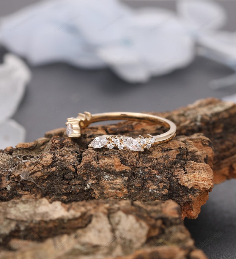 May include: A delicate gold band with a curved row of marquise and round diamonds. The ring is set on a wooden surface.