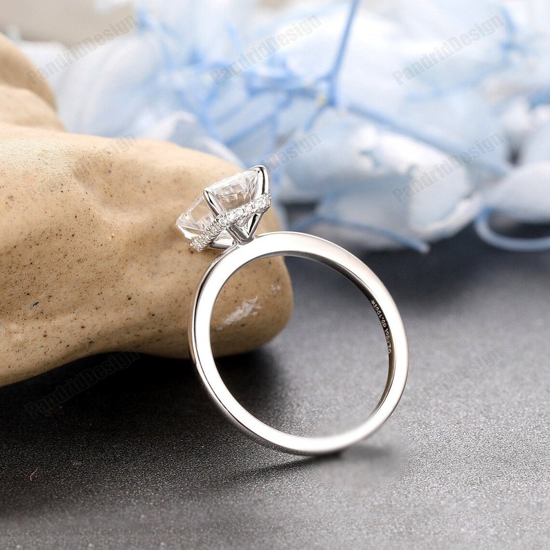 Op de afbeelding: Een zilveren verlovingsring met een grote, vierkant geslepen heldere edelsteen en een halo van kleinere stenen. De ring heeft een gladde, gepolijste band. De ring wordt gepresentteerd op een lichtbruine rots tegen een donkere achtergrond.
