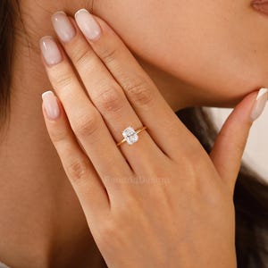 Op de afbeelding: Een gouden ring met een rechthoekige, heldere edelsteen. De ring is gezet op een slanke gouden band. De edelsteen heeft een heldere, rechthoekige vorm. De ring zit aan een vinger.