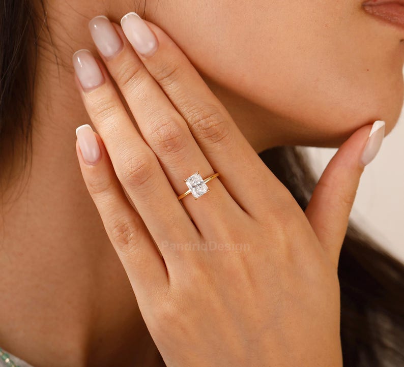 Op de afbeelding: Een gouden ring met een rechthoekige, heldere edelsteen. De ring is gezet op een slanke gouden band. De edelsteen heeft een heldere, rechthoekige vorm. De ring zit aan een vinger.