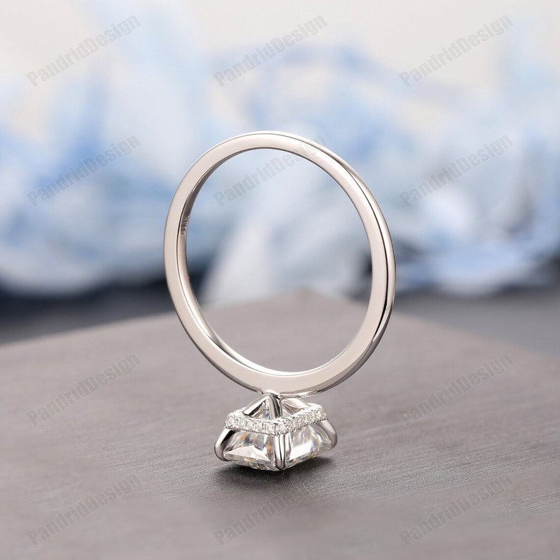 Op de afbeelding: Een zilveren verlovingsring met een vierkant geslepen middensteen en een halo van kleinere stenen. De ringband is dun en gepolijst, en de zetting is gedetailleerd. De ring wordt getoond tegen een vage achtergrond.