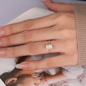 Op de afbeelding: Een gouden ring met een rechthoekige, heldere edelsteen. De ring is gezet op een dunne gouden band. De ring wordt getoond op de vinger van een persoon, met een neutrale achtergrond.