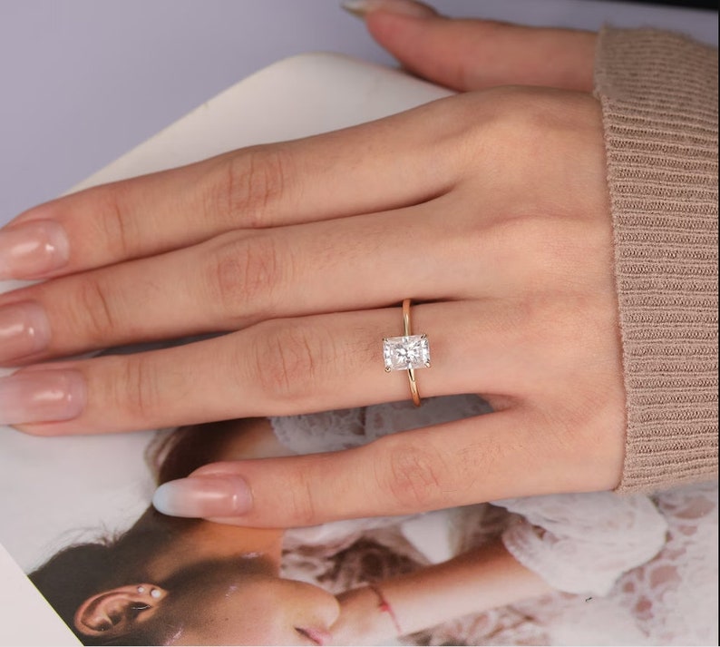 Op de afbeelding: Een gouden ring met een rechthoekige, heldere edelsteen. De ring is gezet op een dunne gouden band. De ring wordt getoond op de vinger van een persoon, met een neutrale achtergrond.