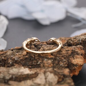 May include: A gold ring with a delicate design featuring small round stones set in a curved band. The ring is shown on a piece of wood.