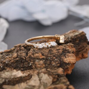 May include: A delicate gold band with a curved row of sparkling marquise-shaped diamonds. The ring is set on a rustic wooden surface.