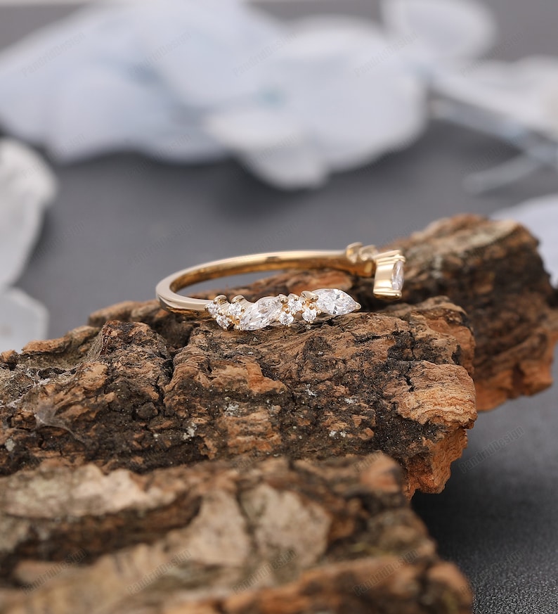 May include: A delicate gold band with a curved row of sparkling marquise-shaped diamonds. The ring is set on a rustic wooden surface.
