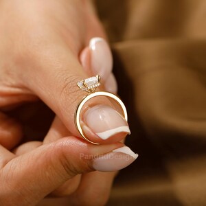 Op de afbeelding: Een gouden ring met een grote, heldere edelsteen en kleinere accentstenen. De ring wordt vastgehouden door de vingers van een persoon, waarbij het ontwerp wordt benadrukt. De band van de ring is van eenvoudig, gepolijst goud. De ring is een sieraad.
