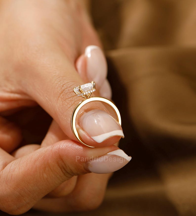 Op de afbeelding: Een gouden ring met een grote, heldere edelsteen en kleinere accentstenen. De ring wordt vastgehouden door de vingers van een persoon, waarbij het ontwerp wordt benadrukt. De band van de ring is van eenvoudig, gepolijst goud. De ring is een sieraad.