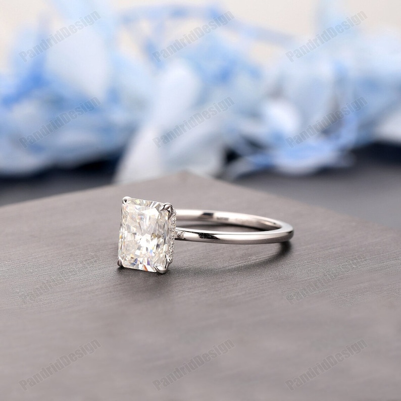 Op de afbeelding: Een verlovingsring met een diamant met een radiant-slijpvorm. De ring heeft een grote, rechthoekige diamant gezet in een delicate, zilveren band. De diamant is het middelpunt en reflecteert het licht tegen een neutrale achtergrond.