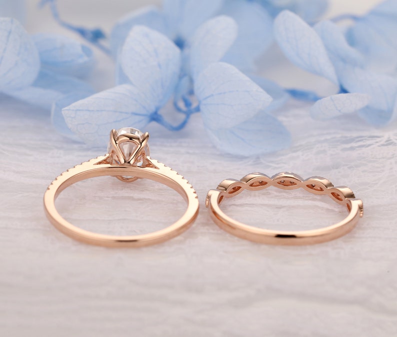 Puede incluir: Un conjunto de anillo de compromiso de oro rosa con una piedra central redonda y una banda a juego con peque&ntilde;as piedras redondas. El anillo est&aacute; sobre un fondo blanco.
