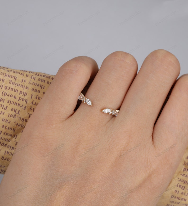May include: A gold ring with a delicate design featuring multiple small, clear gemstones set in a curved shape. The ring is worn on a finger.
