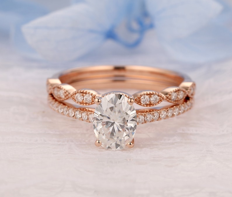 Puede incluir: Un anillo de compromiso de oro rosa con una piedra central ovalada y una banda delicada de piedras m&aacute;s peque&ntilde;as. La banda est&aacute; acentuada con un dise&ntilde;o de grano de mijo.