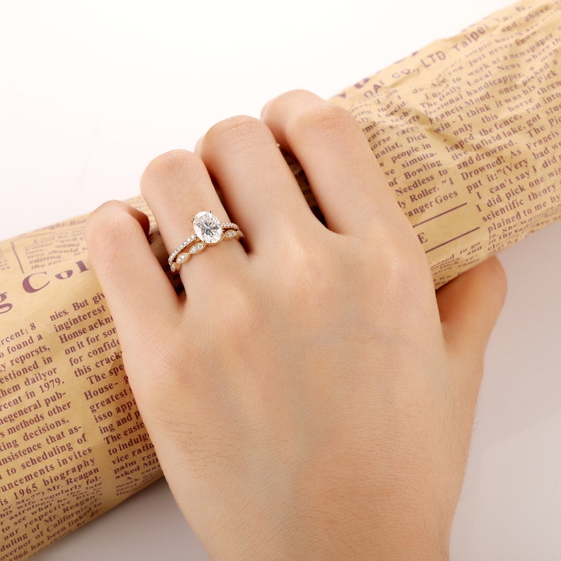 Puede incluir: Un anillo de compromiso de oro de forma ovalada con una banda de peque&ntilde;os diamantes. El anillo est&aacute; en un dedo que lleva una alianza a juego con peque&ntilde;os diamantes.