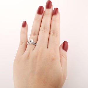 Puede incluir: Primer plano de una mano de mujer con un anillo de compromiso ovalado de plata con una banda de boda a juego. El anillo est&aacute; engastado con peque&ntilde;os diamantes y la banda de boda tiene un dise&ntilde;o delicado.