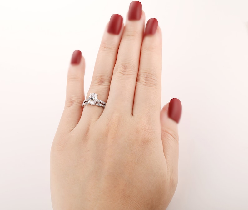 Puede incluir: Primer plano de una mano de mujer con un anillo de compromiso ovalado de plata con una banda de boda a juego. El anillo est&aacute; engastado con peque&ntilde;os diamantes y la banda de boda tiene un dise&ntilde;o delicado.