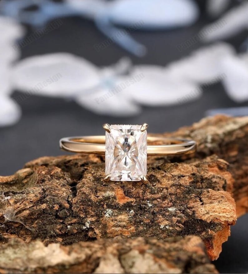 This stunning Hidden Halo Radiant Cut Moissanite Diamond Engagement Ring combines modern sophistication with timeless beauty. The center stone is a radiant cut moissanite, expertly selected for its brilliant sparkle and exquisite clarity. Known for its striking, elongated shape, the radiant cut moissanite is designed to maximize brilliance and fire, creating a captivating focal point.