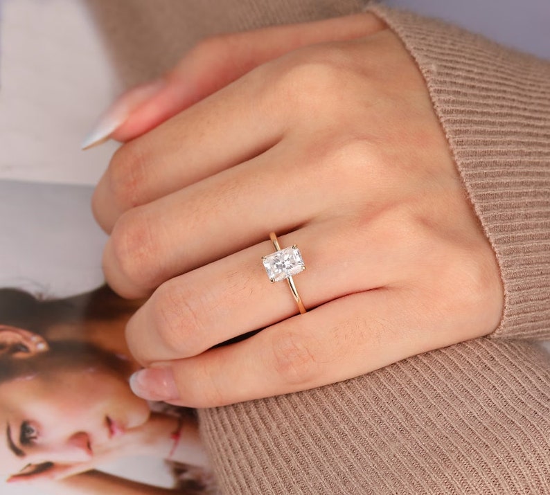 Op de afbeelding: Een gouden ring met een rechthoekige, heldere edelsteen. De ring heeft een slanke gouden band. De edelsteen is gezet in een klassieke vier-klauwzetting. De ring wordt op een vinger getoond.