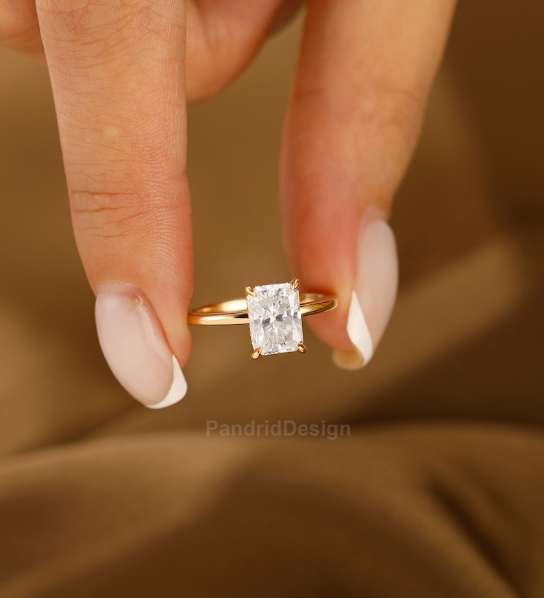 Op de afbeelding: Een gouden ring met een rechthoekige, heldere edelsteen. De ring wordt tussen twee vingers gehouden tegen een bruine achtergrond. De band van de ring is dun en gepolijst. De edelsteen is gezet in een vier-klauwzetting. De tekst "PandridDesign" is zichtbaar.