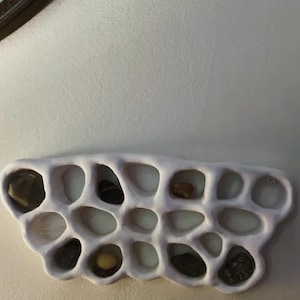 Może przedstawiać: Biała ceramiczna półka ścienna w kształcie plastra miodu. Półka ma 14 przegródek, z których niektóre są wypełnione małymi kamieniami.