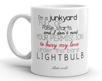 Op de afbeelding: Een witte keramische koffiemok met zwarte tekst die "I'm a junkyard full of false starts and I don't need your permission to bury my love under this bare lightbulb" luidt, waarbij de woorden "bury my love" rood zijn. De tekst "elliott smith" staat in kleinere zwarte tekst onderaan de mok.