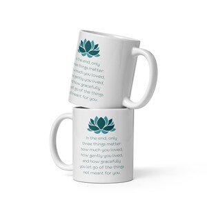Pode incluir: Duas canecas de cerâmica branca com um design de flor de lótus azul-esverdeado e o texto "No final, apenas três coisas importam: o quanto você amou, o quão gentilmente você viveu e o quão graciosamente você deixa ir as coisas que não eram para você."