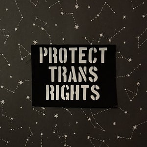 May include: Black fabric patch with white text that reads "PROTECT TRANS RIGHTS" against a dark background with white stars.