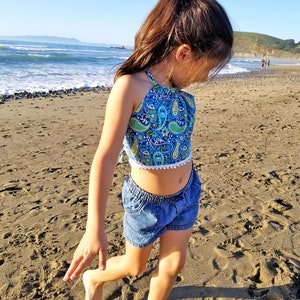 May include: A young person wearing a blue paisley halter top with white trim and denim shorts stands on a sandy beach. The top features a blue and green paisley pattern. The shorts are a light blue denim.