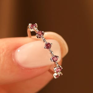 May include: A delicate ring with a series of small, round, pink gemstones set along a thin, silver band. The ring is held between two fingers against a blurred, warm-toned background, highlighting the jewelry's elegant design.