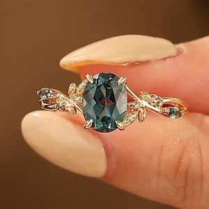 May include: A gold ring featuring an oval, teal-colored gemstone as the centerpiece. The ring has a delicate, leaf-like design with smaller teal stones on either side of the main stone. The ring is held up by a person's hand.