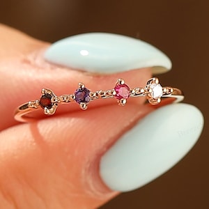 Op de afbeelding: Een delicate gouden ring met vier kleurrijke edelstenen: rood, paars, roze en wit. De edelstenen zijn in een rij langs de band geplaatst. De ring wordt vastgehouden door een persoon met lichtblauwe nagellak.