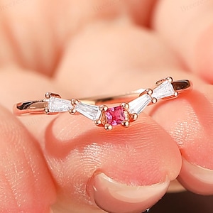 May include: A rose gold ring with a curved design. The ring features a small, round, pink gemstone in the center, flanked by two white baguette-cut stones on each side. The ring is held in a person's hand.