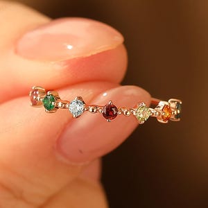 Op de afbeelding: Een delicate gouden ring met een reeks kleurrijke edelstenen in een regenboogopstelling. De edelstenen omvatten groen, blauw, rood en geel, wat een levendige touch toevoegt. De ring wordt door een hand omhoog gehouden.