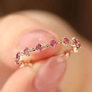 Może przedstawiać: Delikatny złoty pierścionek z ciągłym rzędem małych, żywych różowych kamieni szlachetnych. Obręcz pierścionka jest cienka, polerowana i złota, a kamienie szlachetne są równomiernie rozmieszczone po obwodzie. Pierścionek jest trzymany przez osobę.