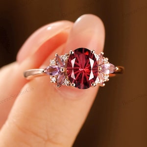 May include: A rose gold ring featuring a large oval-cut, deep red gemstone as the centerpiece. The ring is accented with smaller, light purple gemstones on either side, creating a floral design. The ring is held up against a blurred background.