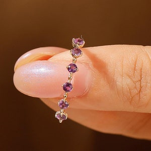 May include: A delicate gold ring with a row of small purple gemstones. The ring is worn on a finger.