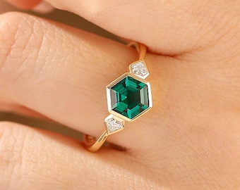 Puede incluir: Un anillo de oro con una piedra preciosa verde hexagonal central flanqueada por dos pequeñas piedras blancas triangulares. La banda es fina y pulida, sujetando las piedras en un engaste de bisel. El diseño resalta la forma geométrica.