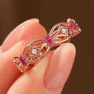 May include: Rose gold ring with a delicate openwork design. The ring features small, rectangular red gemstones and round, clear gemstones. The ring is held in a hand against a blurred background.
