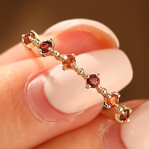 Könnte beinhalten: Ein zarter Goldring mit einer Reihe kleiner, runder, roter und orangefarbener Edelsteine. Der Ring wird vor einem verschwommenen Hintergrund gehalten, der das komplizierte Design und die warmen Farbtöne der Edelsteine zur Geltung bringt.