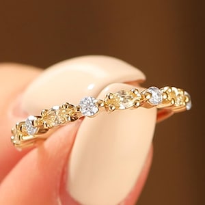 Peut inclure: Une bague en or ornée d'une série de petites pierres précieuses rondes. Alternant des pierres claires et jaunes, la bague est tenue entre les doigts sur un fond flou. Un bijou délicat.