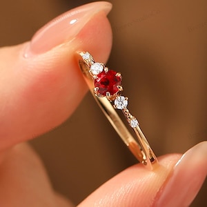 May include: A gold ring featuring a central red gemstone and smaller white gemstones. The ring has a delicate band and is held up by a person's fingers. The ring is a piece of jewelry.