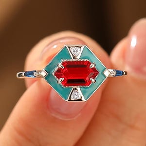 May include: A silver ring with a rectangular red gemstone set within a teal and silver geometric design. The ring features small white accent stones and blue detailing on the band, creating a striking contrast.