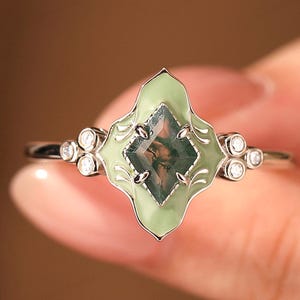 Celebrate your love with this exquisite Handcrafted Mint Green Enamel Engagement Bridal Ring, a stunning piece that combines modern charm with retro elegance. This unique ring features a captivating rhombus-cut 6x8mm real moss agate gemstone, celebrated for its enchanting patterns and vibrant hues of green.