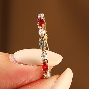 May include: A gold ring with a series of small, colorful gemstones. The ring features red, white, and blue-green stones set in a delicate band. The ring is held between fingers, showcasing its design and sparkle.