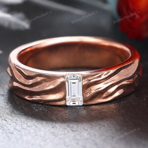 May include: Rose gold ring featuring a textured band and a clear, rectangular gemstone. The ring is set against a dark background with a blurred floral element. The band has a wave-like pattern, adding a unique design element.