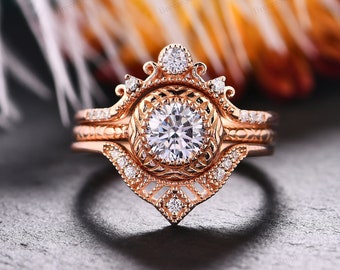 Può includere: Un anello di fidanzamento in oro rosa con una grande pietra centrale rotonda e pietre di accento più piccole. L'anello presenta un design delicato e intricato con una parte superiore a forma di corona e una fascia con un motivo testurizzato.