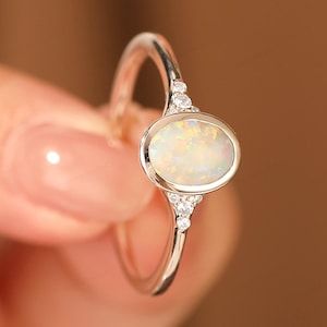 May include: A silver ring featuring an oval opal gemstone. The opal is surrounded by a silver setting and accented with small, clear gemstones on each side. The ring is held up against a blurred background.