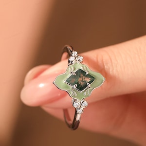 Puede incluir: Anillo de plata con un engaste en forma de hoja verde claro y una piedra cuadrada verde musgo. Peque&ntilde;as piedras redondas y claras est&aacute;n engastadas a cada lado del engaste. Una joya.