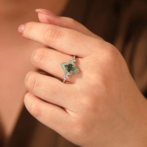 Puede incluir: Un anillo de plata con un dise&ntilde;o de tr&eacute;bol de cuatro hojas, con una piedra preciosa verde claro en el centro y peque&ntilde;as piedras claras. El anillo se lleva en una mano, mostrando sus delicados detalles y su elegante estilo.
